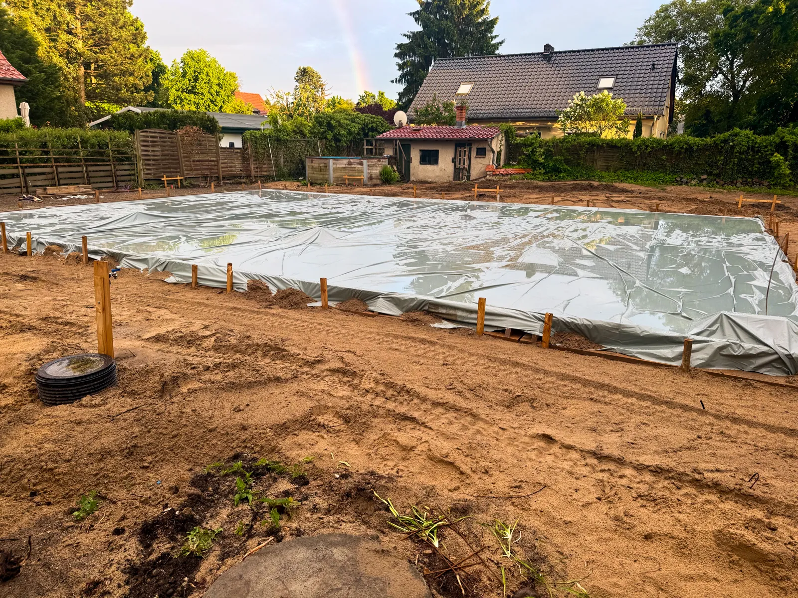 Frisch betonierte Bodenplatte auf einer Baustelle, vollständig mit einer Folie gegen Regen abgedeckt und an den Rändern mit Schalhölzern gesichert — im Hintergrund ein Regenbogen nach dem Schauer