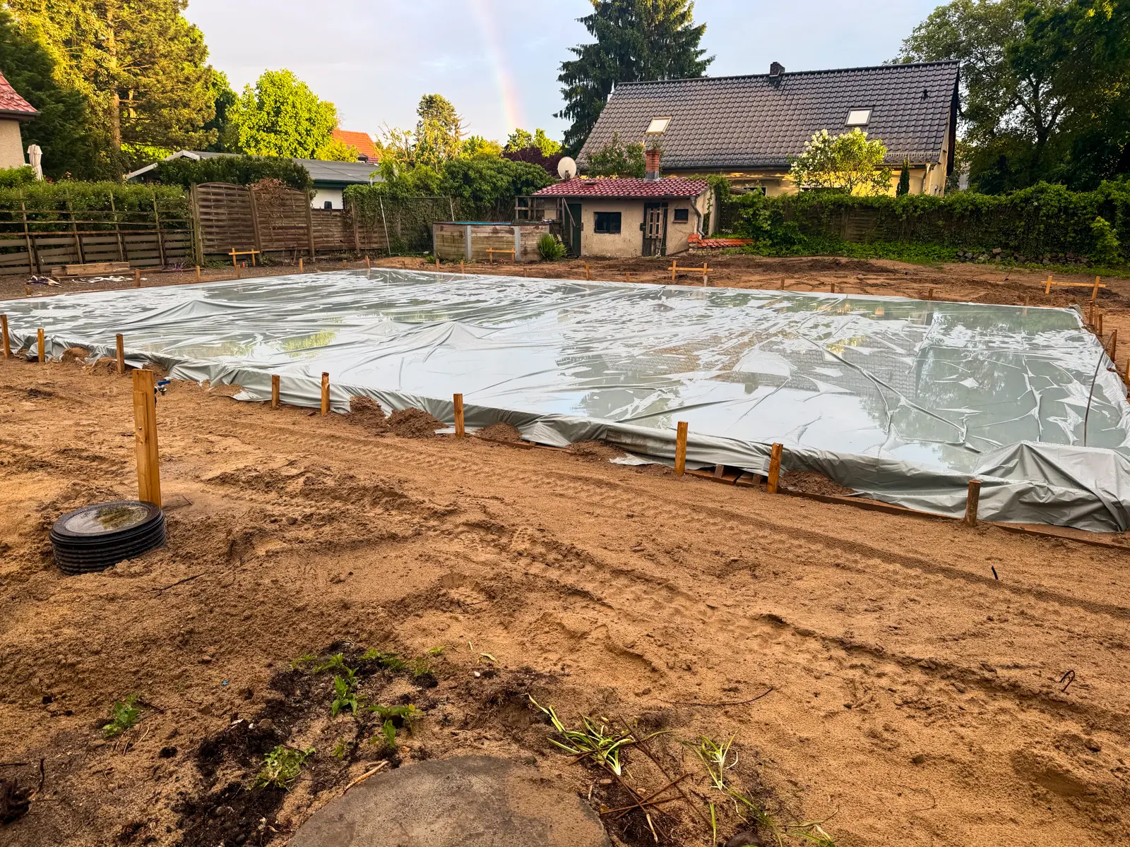 Frisch betonierte Bodenplatte auf einer Baustelle, vollständig mit einer Folie gegen Regen abgedeckt und an den Rändern gesichert — im Hintergrund ein Regenbogen nach dem Schauer